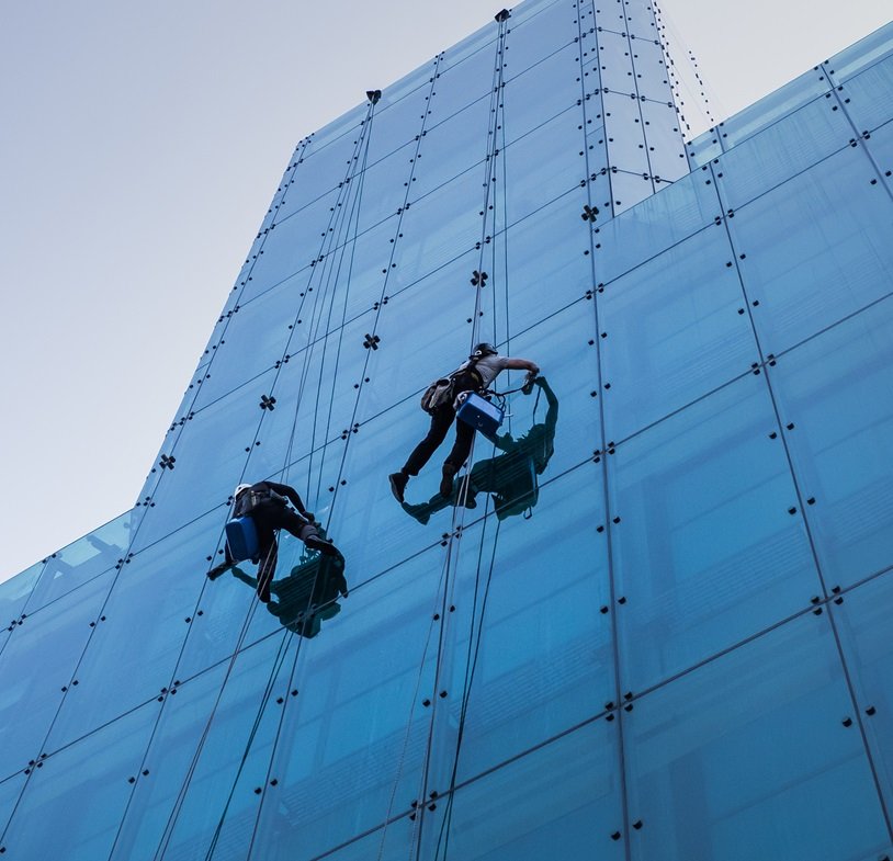 Vertical low angle shot of two people climbing a tall glass building during the day time A vertical low angle shot of two people climbing a tall glass building during the day time